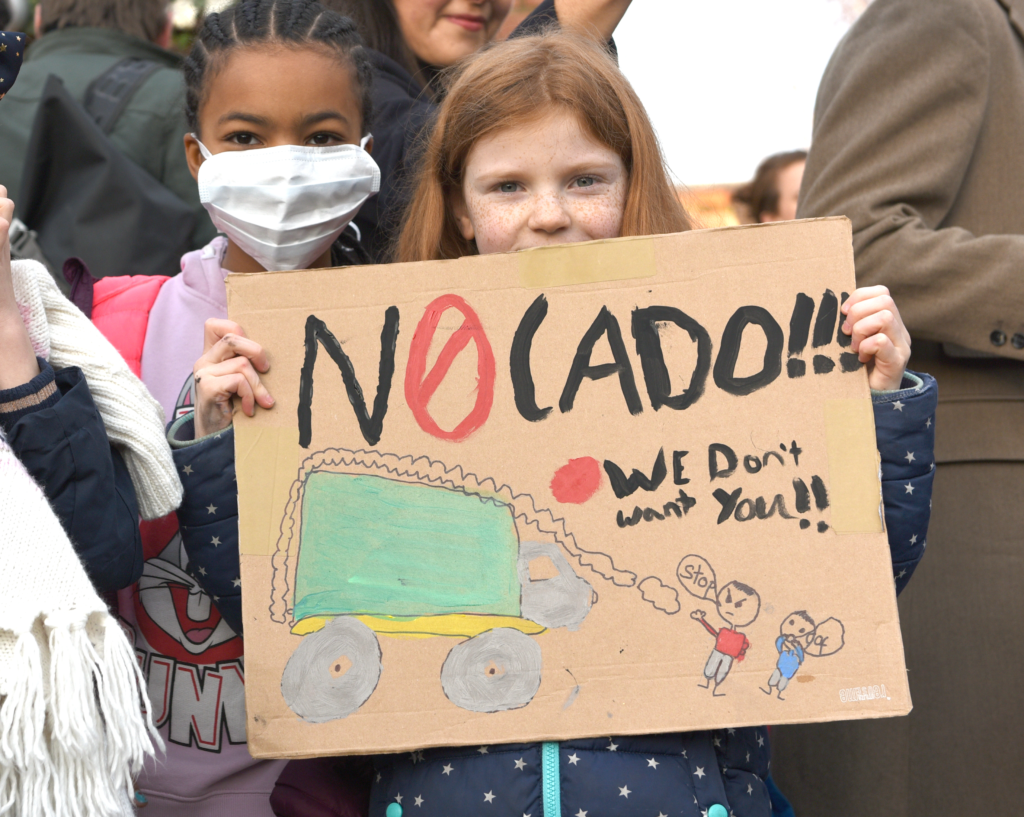 Two kids holding a sign saying NOcado with a painting of a polluting van and two kids saying stop we don't want you!!