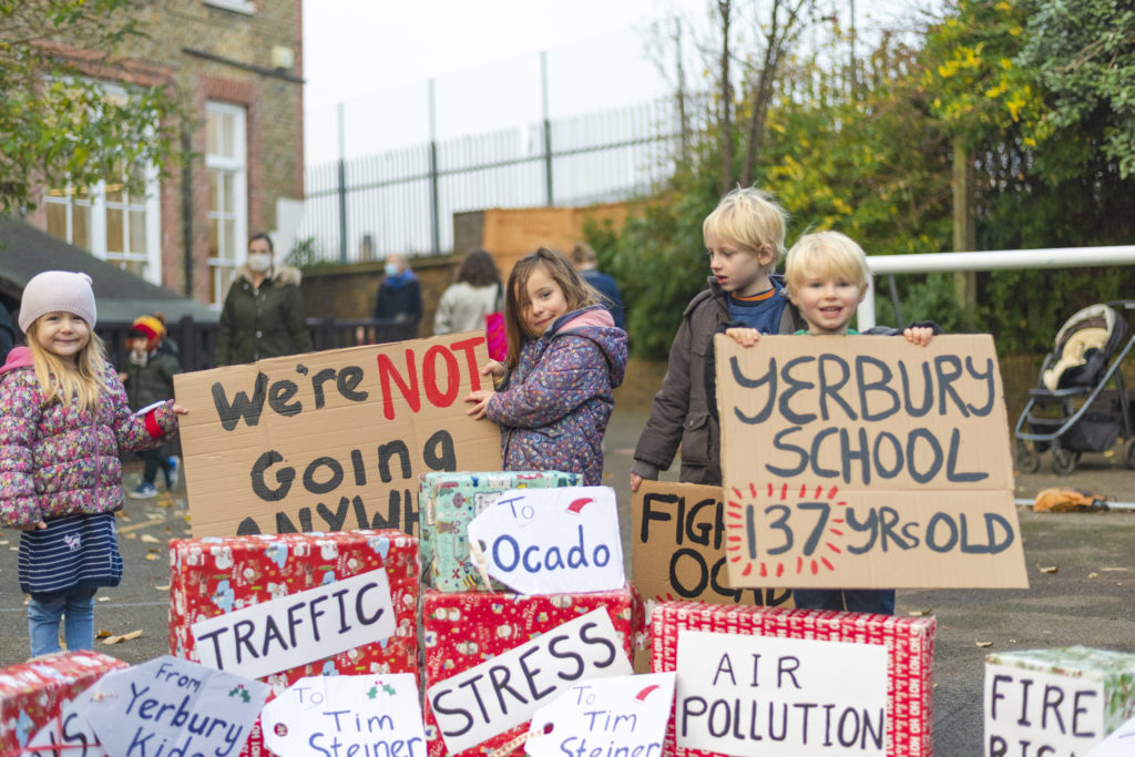 Nursery kids standing in a pile of christmas presents labelled air pollution, traffic, stress, fire risk and holding signs saying Yerbury School 137 yrs old and We're not going anywhere